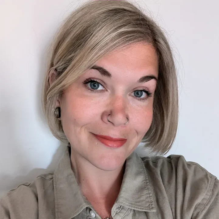 A person with short, light blonde hair and blue eyes smiles at the camera. They are wearing a beige collared shirt, hoop earrings, and have a nose ring. The background is plain and light-colored.