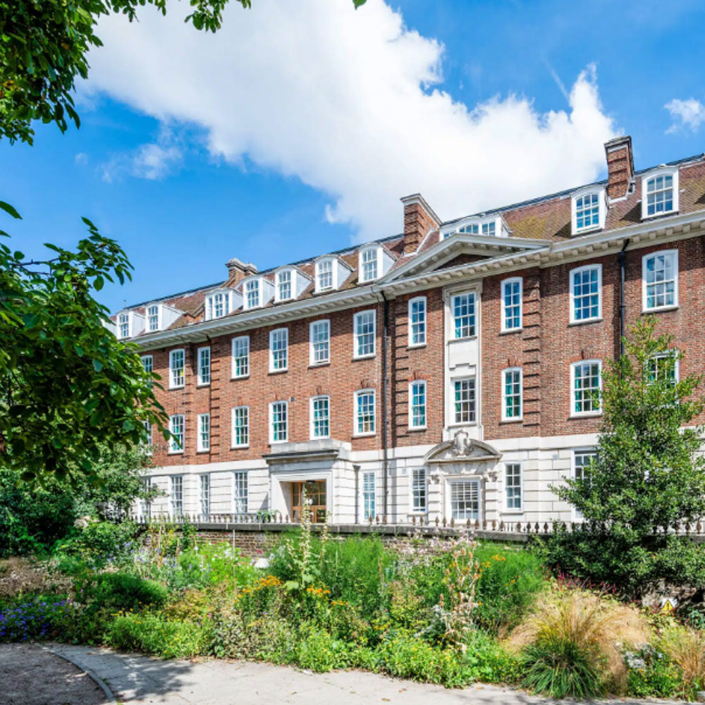 A large red-brick accommodation with white-framed windows and a main entrance, surrounded by green trees, shrubs, and a flower garden under a blue sky with scattered clouds.
