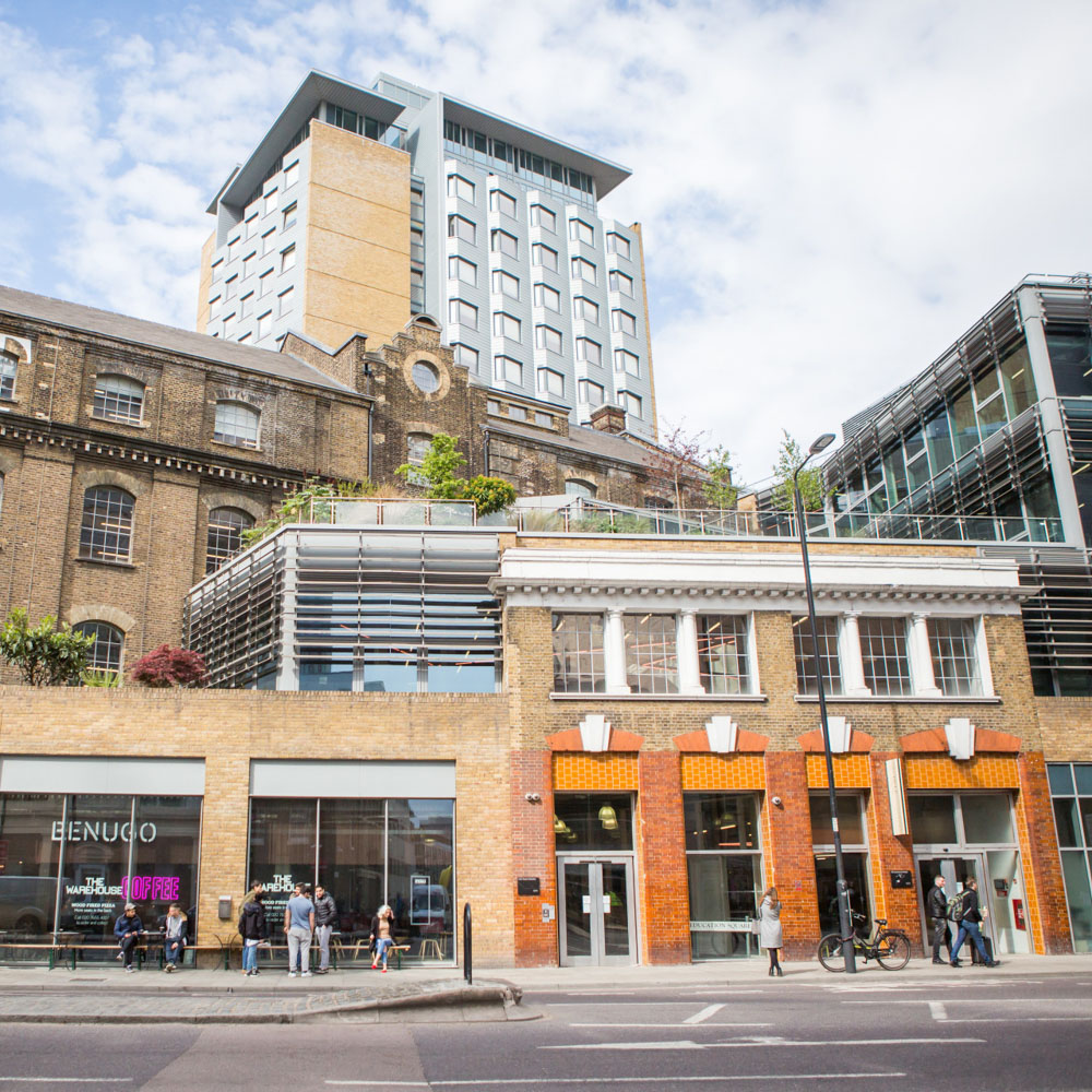 A city street view shows a mix of modern and historic buildings, with people walking and a few bicycles parked. The sky is partly cloudy, and greenery is visible on a terrace above stylish accommodation along the street.