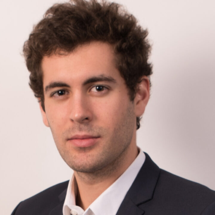 A young man with curly brown hair wearing a dark suit jacket and white shirt poses against a plain light background, looking confidently at the camera.