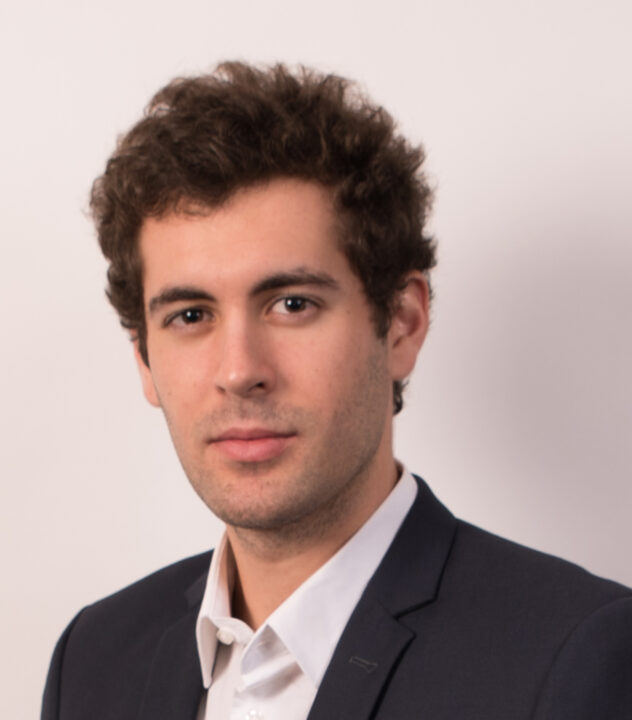 Clement Ruah, a young man with curly brown hair, wearing a dark suit jacket and a white dress shirt, poses against a plain light background, looking confidently at the camera.
