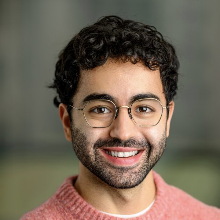 A young man with curly dark hair, glasses, and a beard smiles at the camera. He is wearing a pink textured sweater and standing against a softly blurred, neutral background.