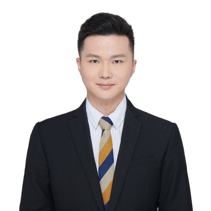 A young man with short black hair wearing a black suit jacket, white and blue striped shirt, and a yellow, blue, and white striped tie, posing and smiling slightly in front of a plain white background.