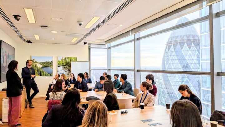 A group of people listen to two presenters in a modern meeting room with large windows, overlooking the Gherkin building in London. Some attendees are seated at tables, taking notes or holding coffee cups.