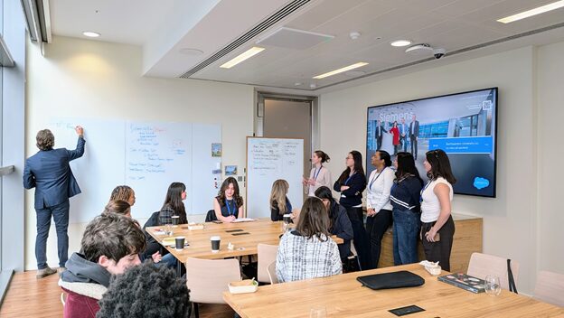 A group of people in a modern meeting room; one person writes on a whiteboard while others stand and watch. Several people sit at tables with drinks and notepads, and a large screen displays a presentation.