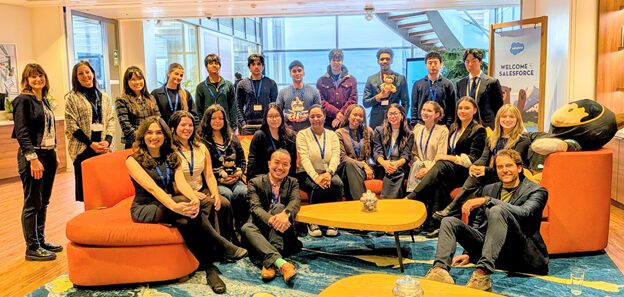 A diverse group of people, mostly young adults, pose and smile in a bright, modern office lounge with orange seating, a blue rug, and Salesforce branding in the background.