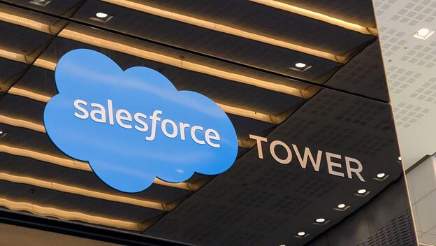A blue Salesforce cloud logo and the word “TOWER” are displayed on a modern building’s exterior, with reflective panels and ceiling lights visible above the sign.