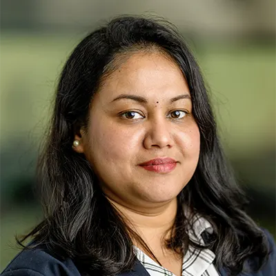 Rashi Dutt, with long black hair, wearing a dark blazer and a white patterned shirt, looks at the camera with a slight smile against a blurred background.