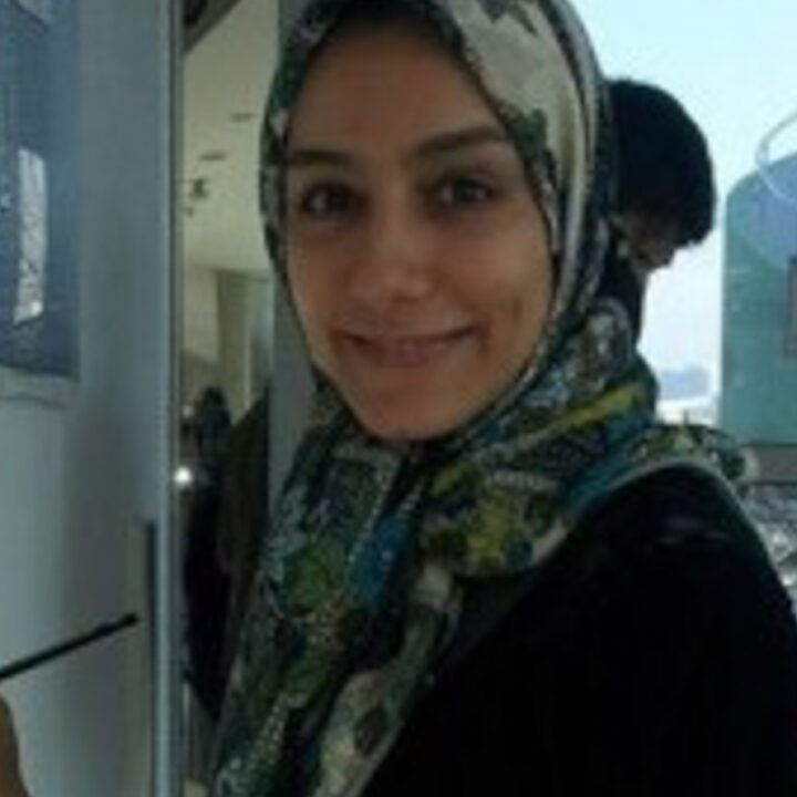A woman wearing a patterned headscarf smiles at the camera while standing indoors near a window.