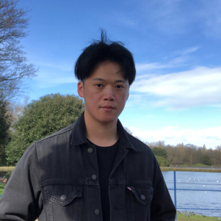 A young man with short black hair wearing a black denim jacket stands outdoors near a body of water, with trees and a blue sky in the background.