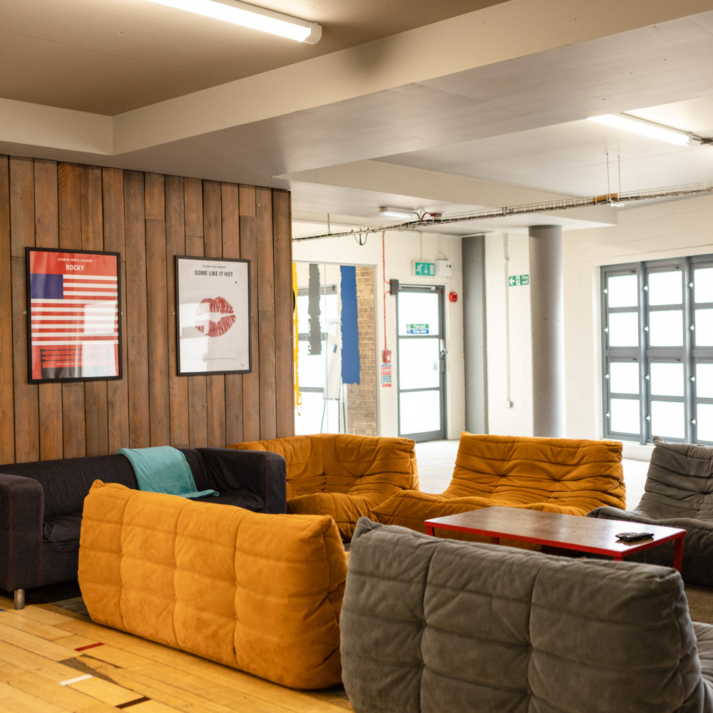 A modern accommodation lounge area with mustard yellow and gray sofas arranged around a red coffee table, wooden wall panels displaying framed posters, and large windows letting in natural light.
