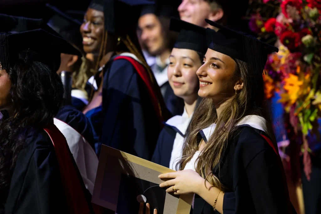 A group of graduates wearing caps and gowns smile and look ahead during a graduation ceremony. One graduate in the foreground holds a diploma cover, while colorful flowers brighten the background—perfect for any Graduation FAQ visuals.