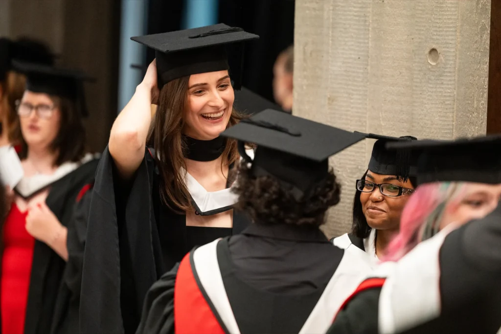 A group of graduates in caps and gowns smile and talk together indoors. One woman in the center adjusts her cap and laughs while others stand nearby, discussing plans and a Graduation FAQ as they prepare for the ceremony.