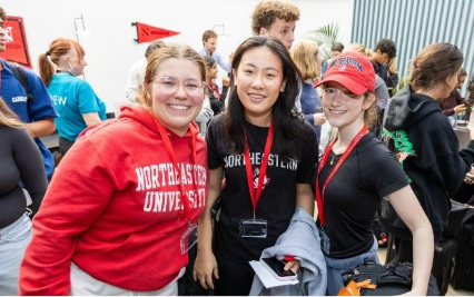 Three young women wearing Northeastern University lanyards smile and pose together at a busy indoor event, possibly discussing campus accommodation options. One wears a red sweatshirt, another a black shirt, and the third a black cap.