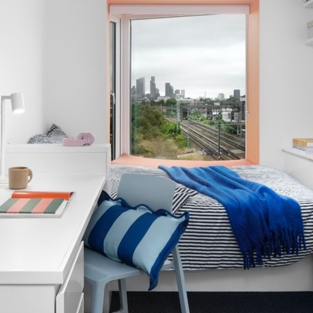 A small, modern accommodation featuring a desk, chair, and bed by a large window. The bed has blue-striped bedding and a blue throw. City buildings and train tracks are visible outside under a cloudy sky.