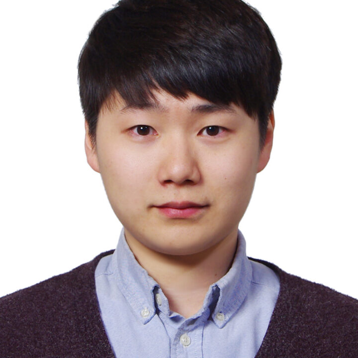A young person with short, dark hair, wearing a light blue button-up shirt and a dark sweater, poses for a headshot against a plain white background.