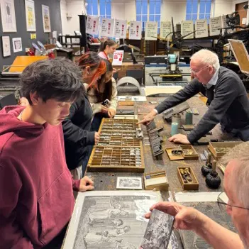 A group of people gathered around a table covered with printing materials and tools in a workshop, with one person showing a print while others examine type cases and printing blocks.