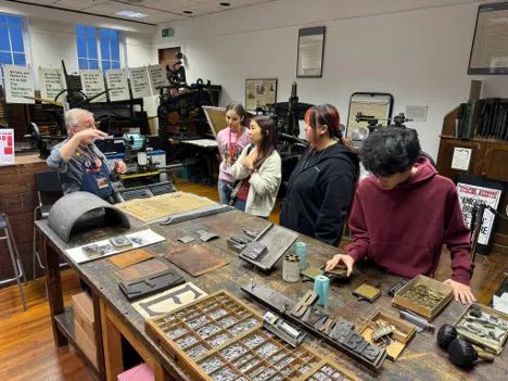 A group of five people stands around a large table covered with printing tools and type blocks in a printmaking studio while one person gestures and explains to the others.