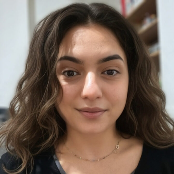 A young woman with wavy brown hair and natural makeup smiles slightly at the camera. She is indoors, with shelves and books blurred in the background.