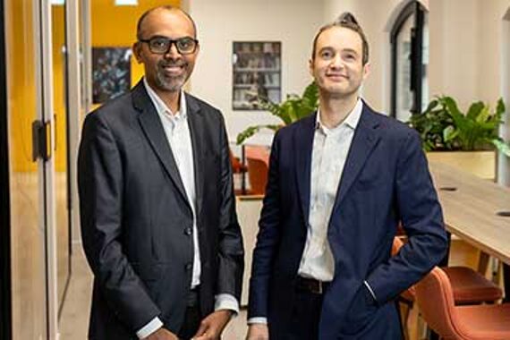 Two men in business suits stand and smile in a modern office space with plants, orange chairs, and large windows in the background.