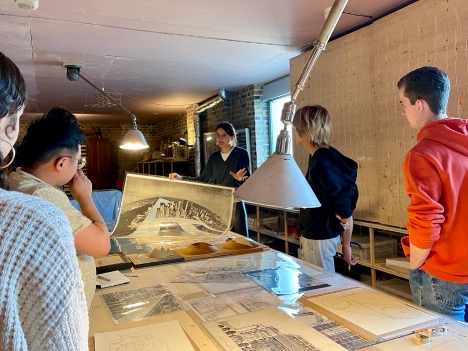 A group of people stand around a table covered with architectural drawings as a woman holds up a large print and speaks, in a room with brick walls and overhead lamps.