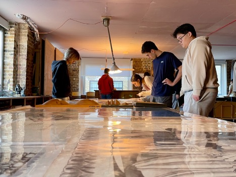 Four people stand around a large table covered with papers or artwork in a well-lit room with brick walls and large windows, intently examining the materials.