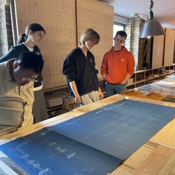 Four people stand around a table looking at large blueprints and architectural drawings in a well-lit room with exposed brick walls and shelves in the background.