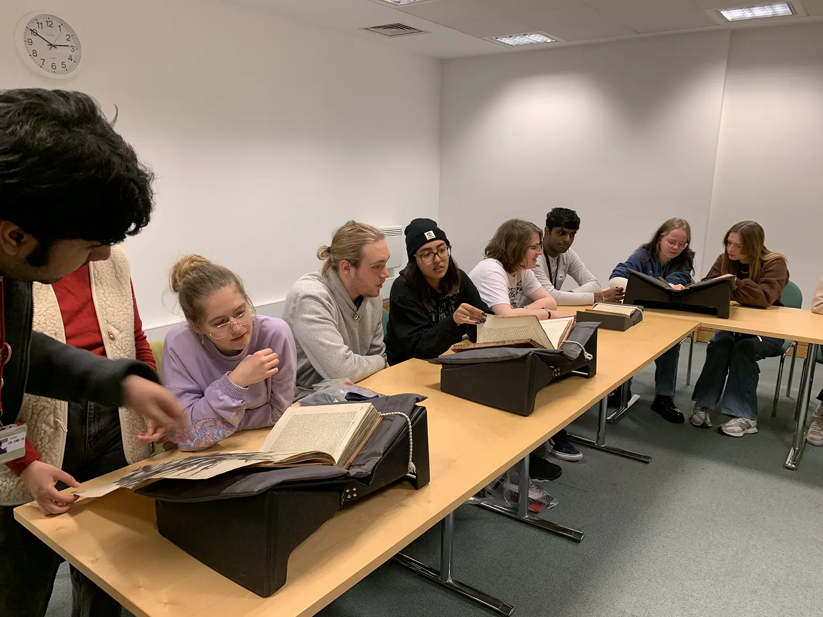 A group of people sitting at a long table in a classroom, examining old books placed on book supports, with some individuals discussing and others reading. A clock and a whiteboard are visible on the wall.