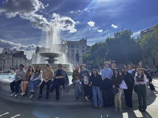 A group of people sit and stand along the edge of a large fountain in a city square on a sunny day. Water sprays from the fountain, and historic buildings and trees are visible in the background under a partly cloudy sky.