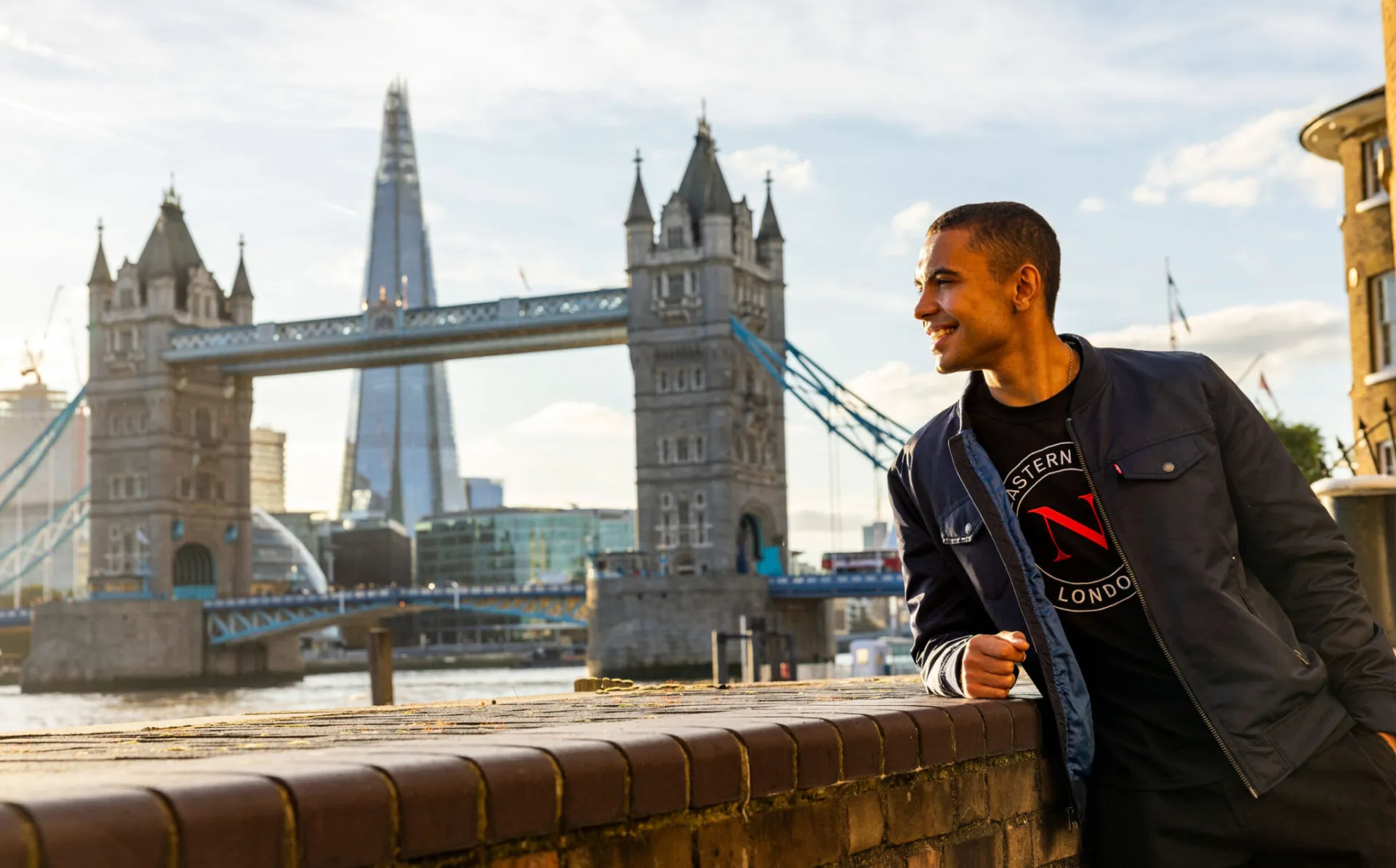 A man smiles while leaning on a brick wall beside the River Thames, with Tower Bridge, The Shard skyscraper, and a nearby university in the background on a sunny day in London.