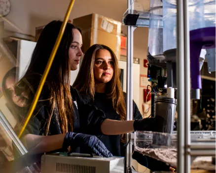 Two women wearing gloves work with scientific equipment in a university lab, focusing intently on a large, clear machine with tubes and containers amid an array of machines, wires, and laboratory tools.