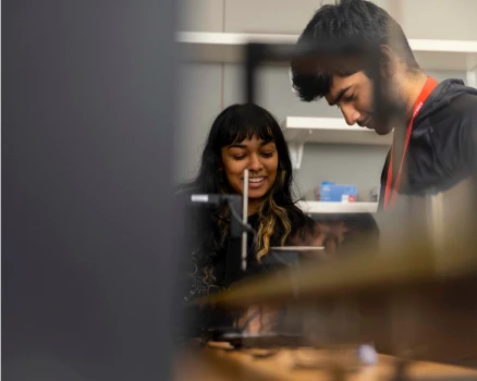 Two people stand closely together in a university lab setting, focusing on a project. One is smiling while the other examines equipment, with shelves and tools visible in the background.