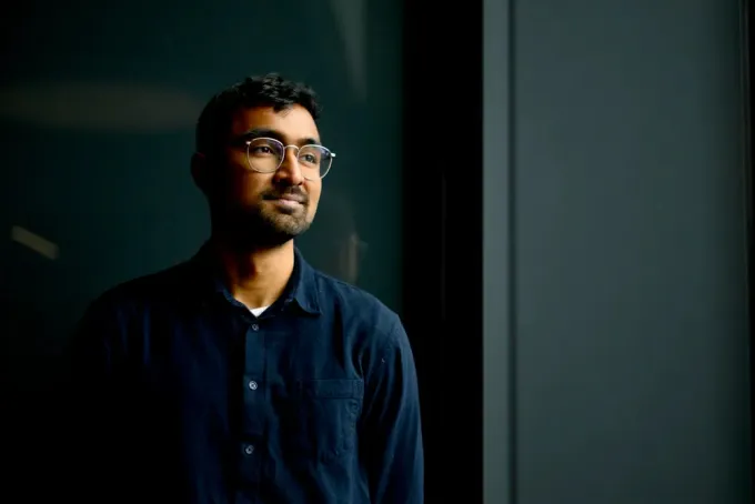 A man wearing glasses and a dark button-up shirt stands indoors, gazing thoughtfully out of the frame, as if reflecting on his day at university. The background is dark, with light softly illuminating one side of his face.