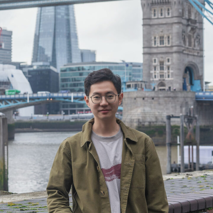 A young man wearing glasses and an olive green jacket stands and smiles in front of the River Thames, with Tower Bridge and modern London buildings visible in the background.