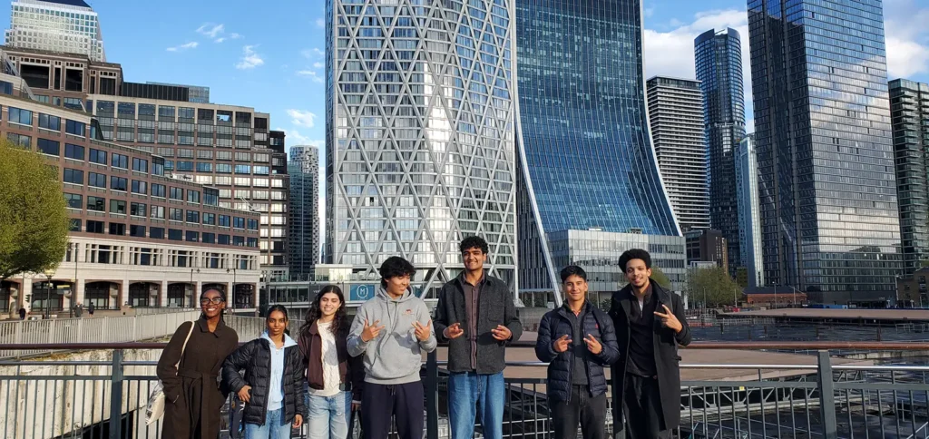 A group of seven people pose and smile on a walkway with modern skyscrapers and office buildings in the background on a sunny day.
