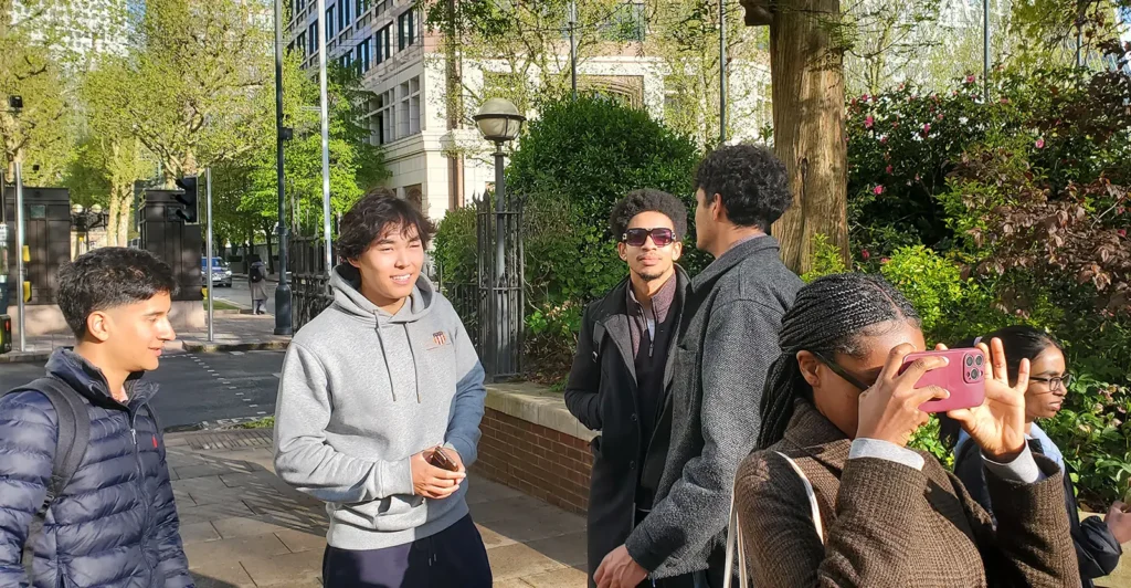 A group of young adults stands outdoors on a sunny day in a city. One person is taking a photo with a pink phone while others smile and talk casually in front of trees and buildings.
