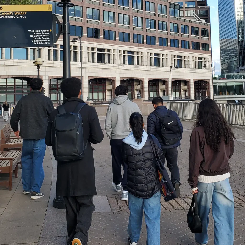 A group of six people walk together on a paved path in an urban area near benches and a building. Most are wearing jackets and carrying bags. A yellow sign for Canary Wharf is visible above them.