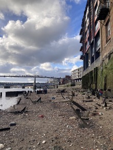 A cloudy sky over a rocky, debris-strewn riverbank beside tall buildings, with a bridge in the background and a few people walking along the shore.