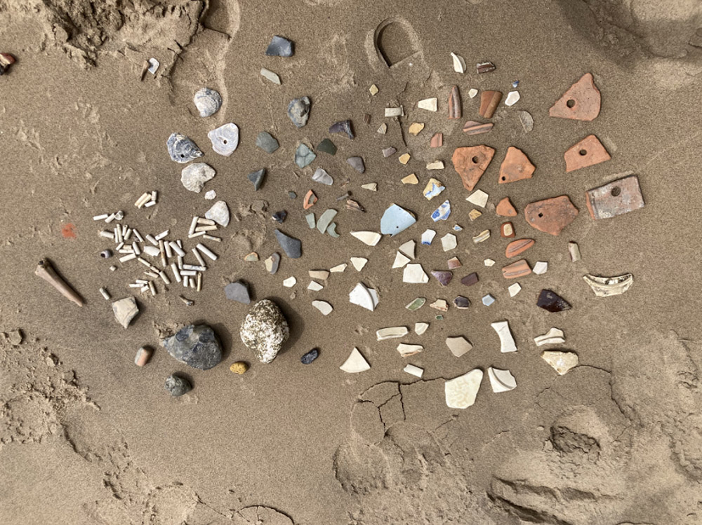 Assorted small rocks, pottery shards, shells, and beach debris are arranged in neat rows and clusters on wet sand, creating a colorful, organized display of natural and manmade objects.