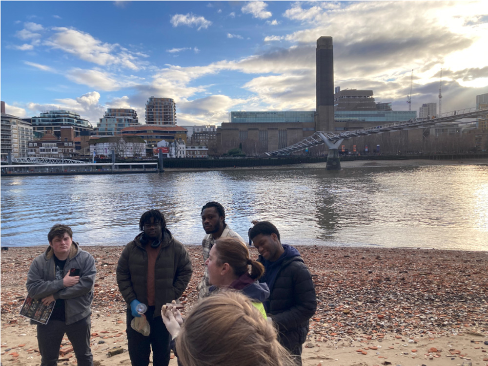 A group of people stand on a pebbled beach by the Thames River, with London's Millennium Bridge and Tate Modern visible across the water under a partly cloudy sky.