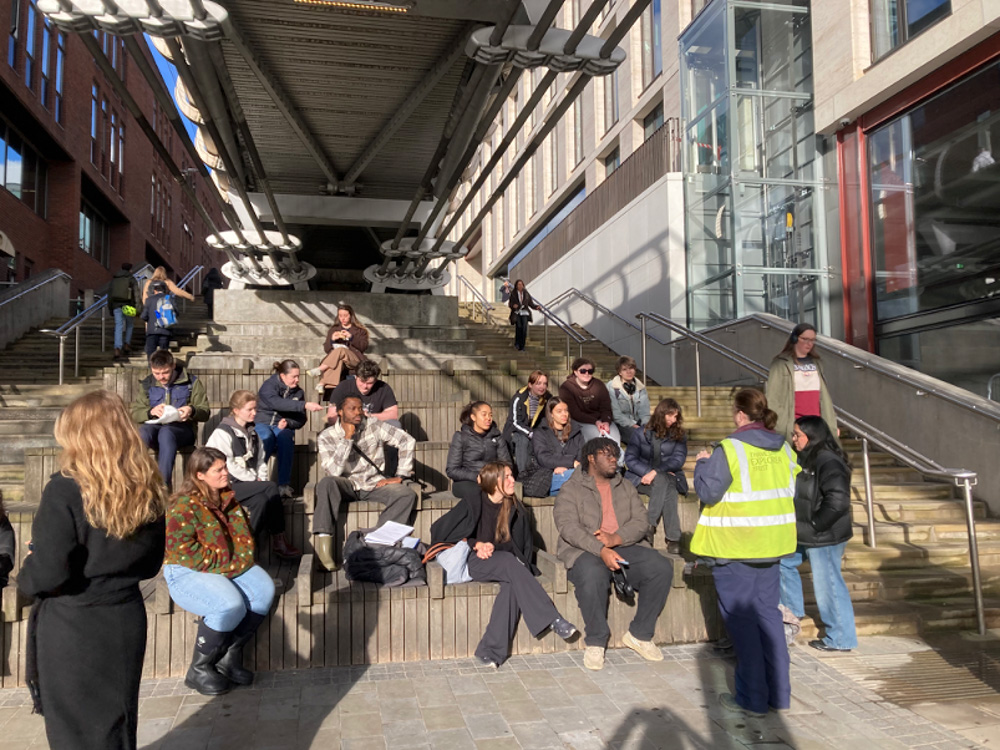 A group of people sit and stand on outdoor steps in the sunlight near a modern building; one person in a yellow hi-vis vest addresses the group while others listen or talk among themselves.