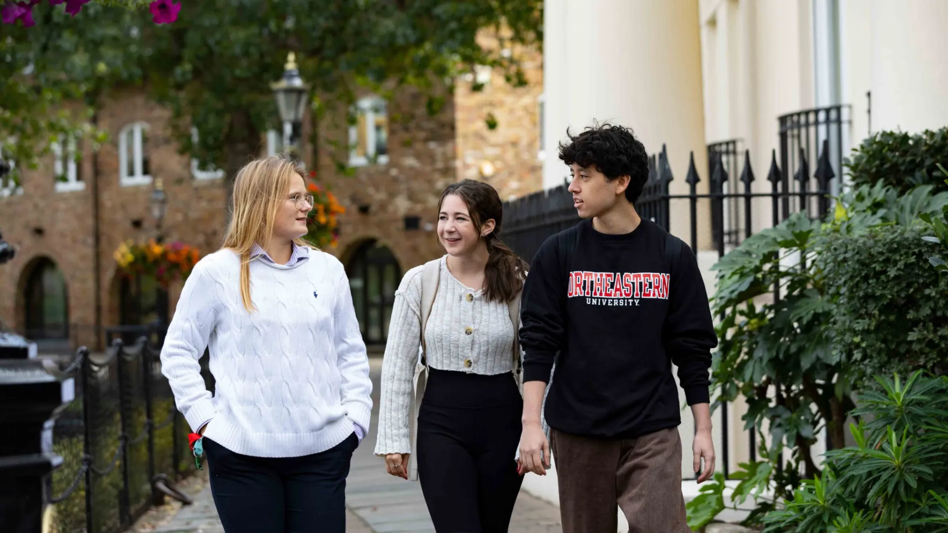Three young adults walk together outside on a sidewalk, smiling and talking. One wears a white sweater, another a cream cardigan, and the third a Northeastern University sweatshirt, surrounded by greenery and university buildings.