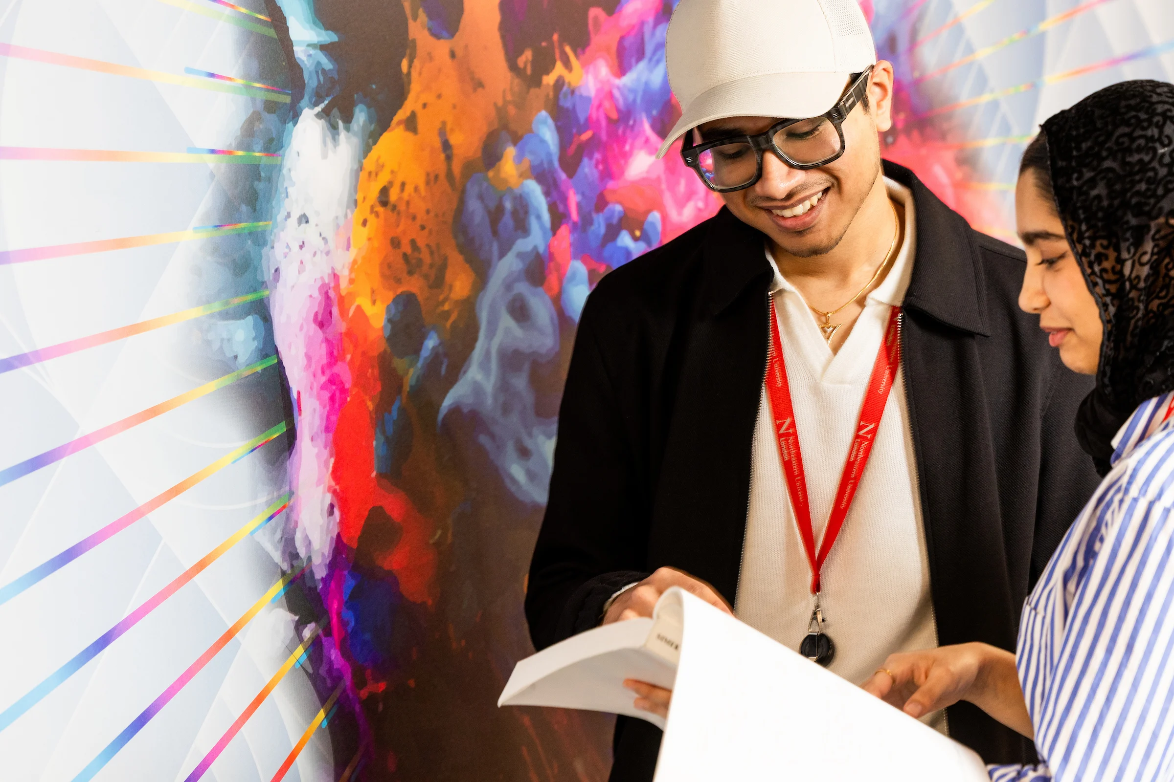 Two people stand in front of a colorful abstract mural, smiling and looking at open books or papers together. In the clearing of lively colors, one wears a cap and glasses; the other wears a striped shirt and a headscarf.