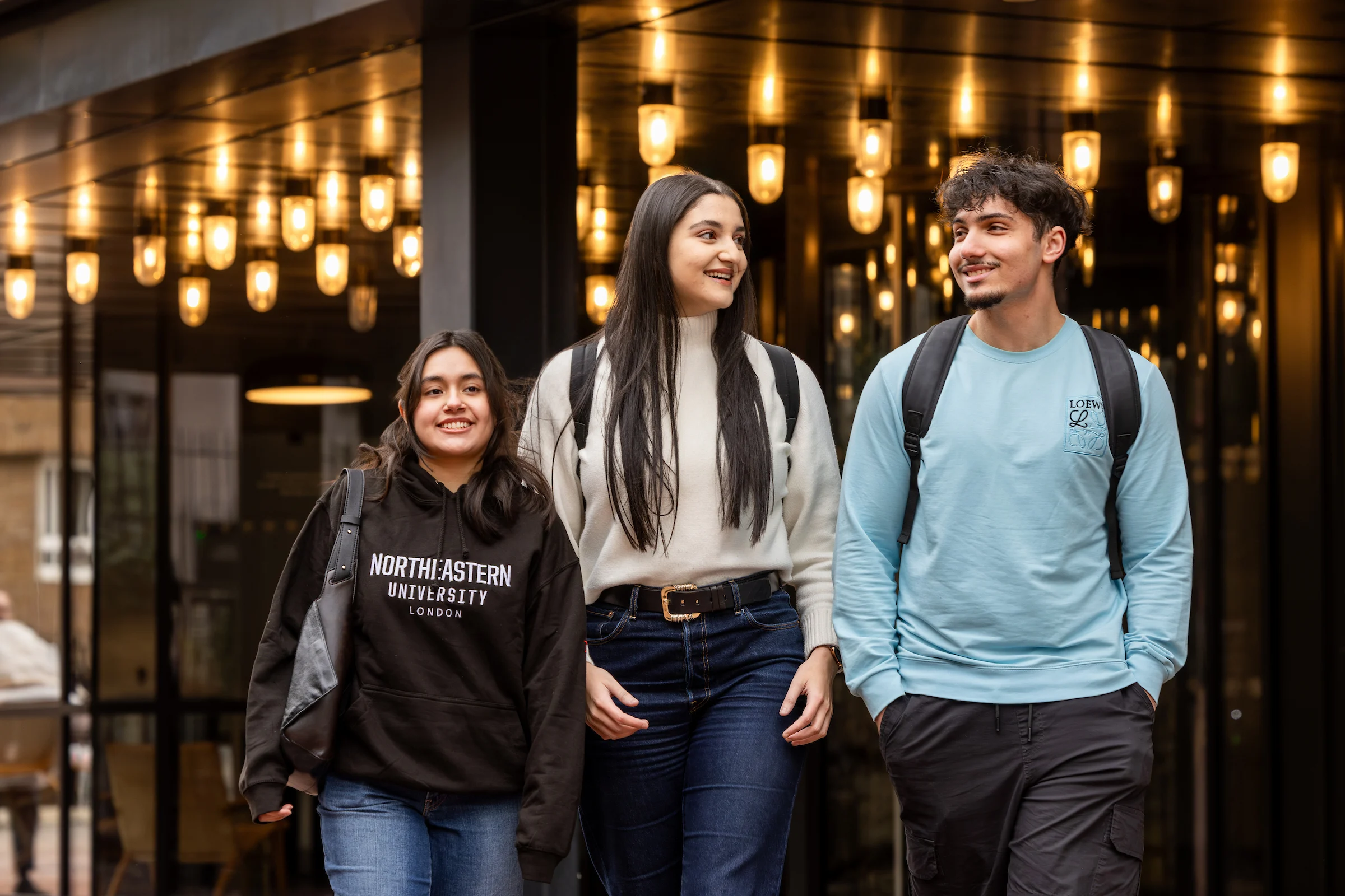Three smiling young adults walk together outside a building with hanging lights. Dressed casually with backpacks—one in a Northeastern University London hoodie—they discuss university life and opportunities through clearing.