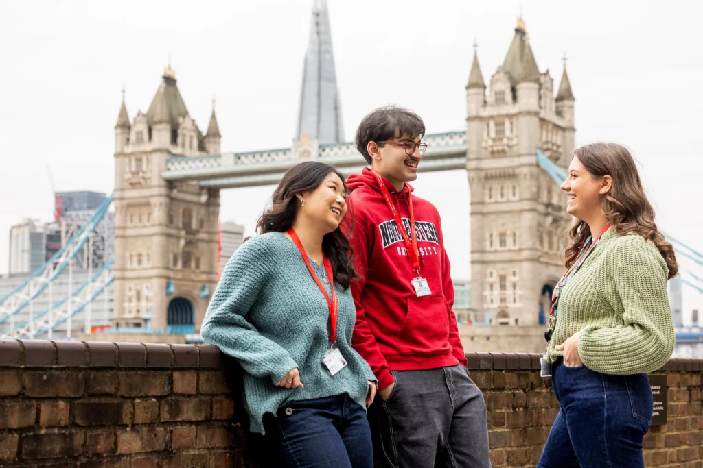 Three young adults, two women and one man, stand and chat by a brick wall with Tower Bridge and The Shard in the background. Wearing casual clothes and university clearing lanyards, they smile and laugh together.