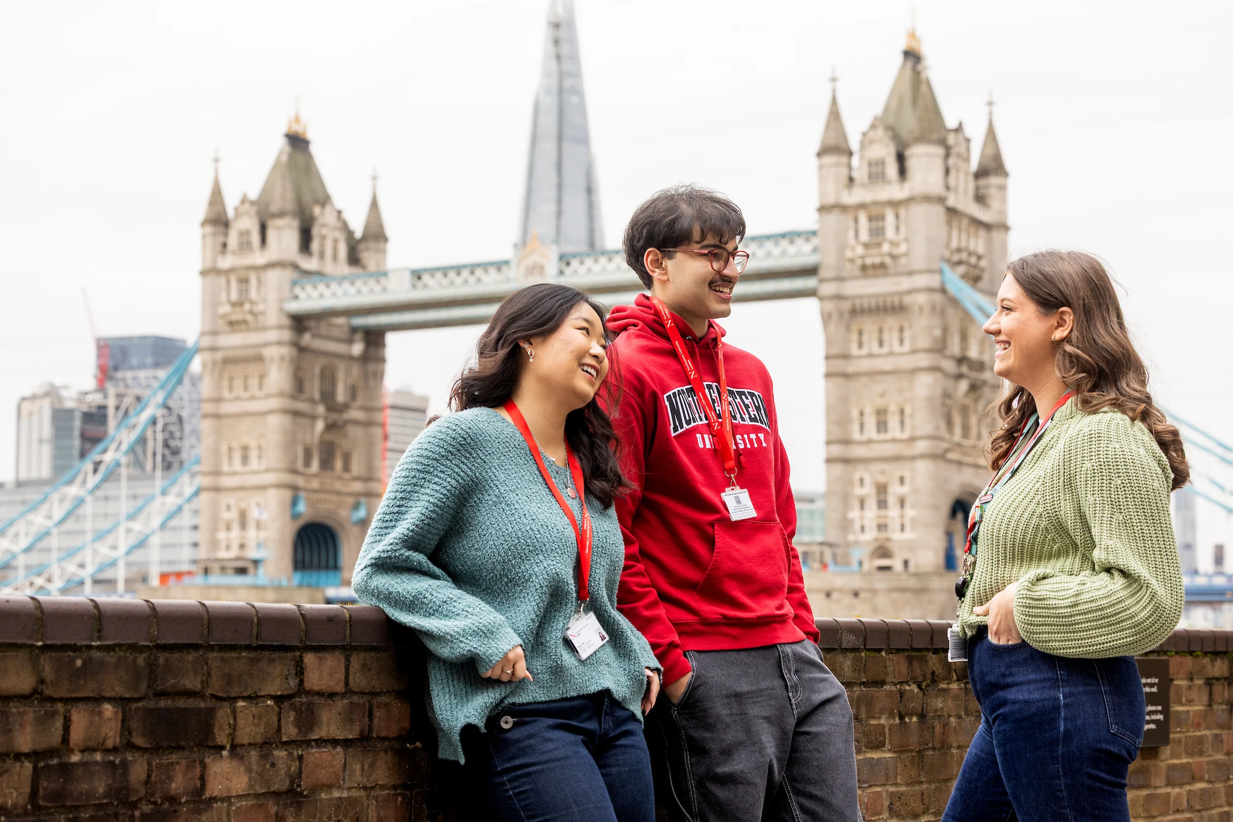 Three young adults, two women and one man, stand and chat by a brick wall with Tower Bridge and The Shard in the background. Wearing casual clothes and university clearing lanyards, they smile and laugh together.