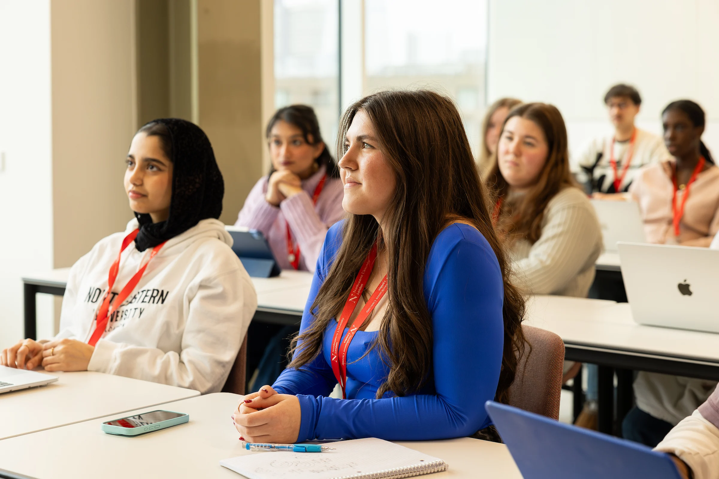 A group of students, wearing red lanyards, sit attentively at desks in a bright classroom during a clearing session. Some have laptops and notebooks open as they listen to a lecture or presentation.