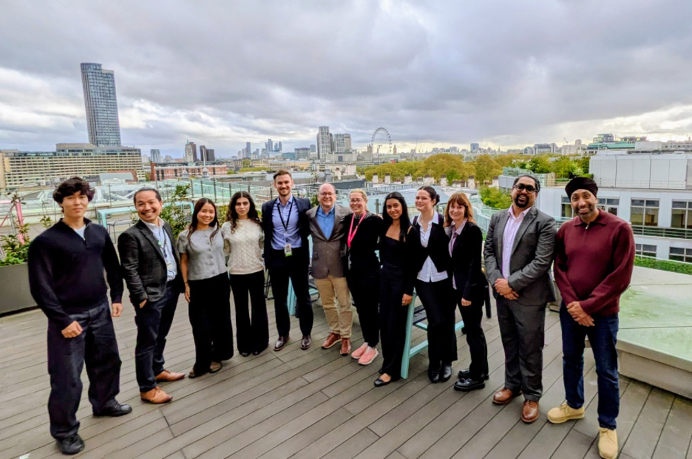 A group of thirteen people pose together on a rooftop deck with a city skyline and the London Eye visible in the background under a cloudy sky.