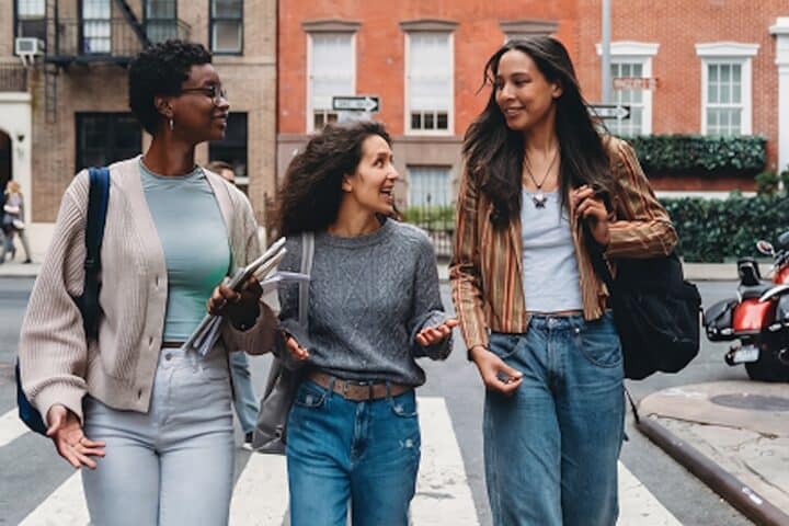 Three young women walk together on a city street, smiling and talking. They wear casual clothes, carrying bags and notebooks, and appear engaged in friendly conversation. Brick buildings and a red motorcycle are in the background.