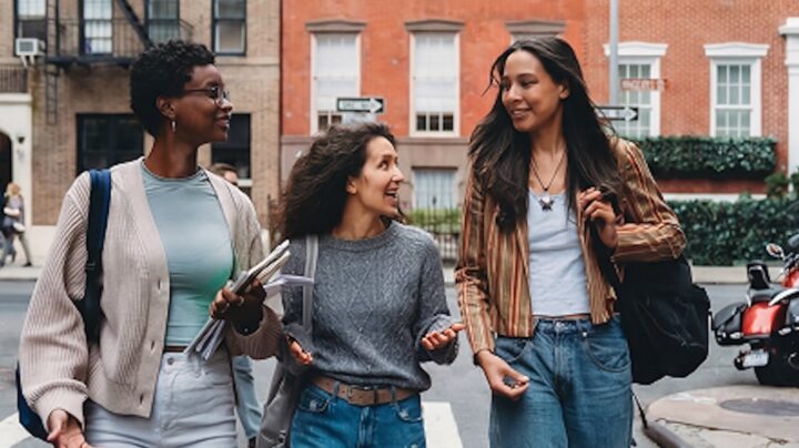 Three young women walk together on a city street, smiling and talking. They wear casual clothes, carrying bags and notebooks, and appear engaged in friendly conversation. Brick buildings and a red motorcycle are in the background.